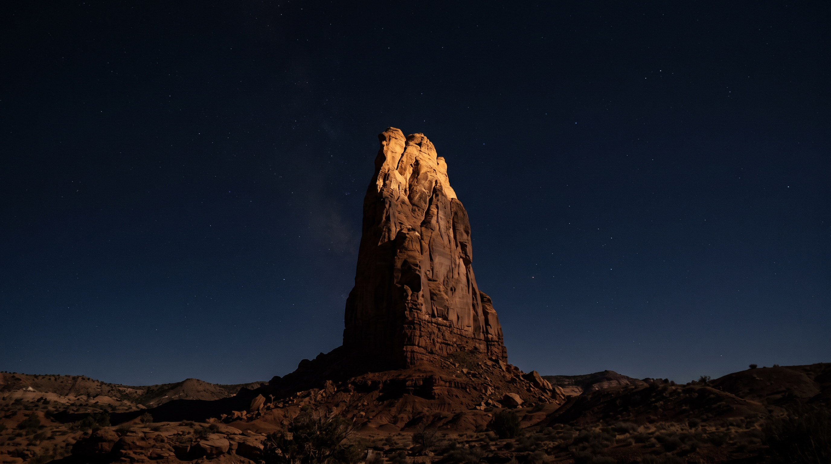 A monolithic sandstone spire lit by warm light against a starlit navy night sky